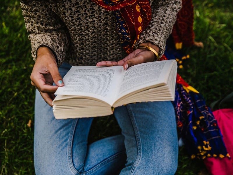 A person seated on grass, wearing a beige sweater and blue jeans, is holding and reading an open book.