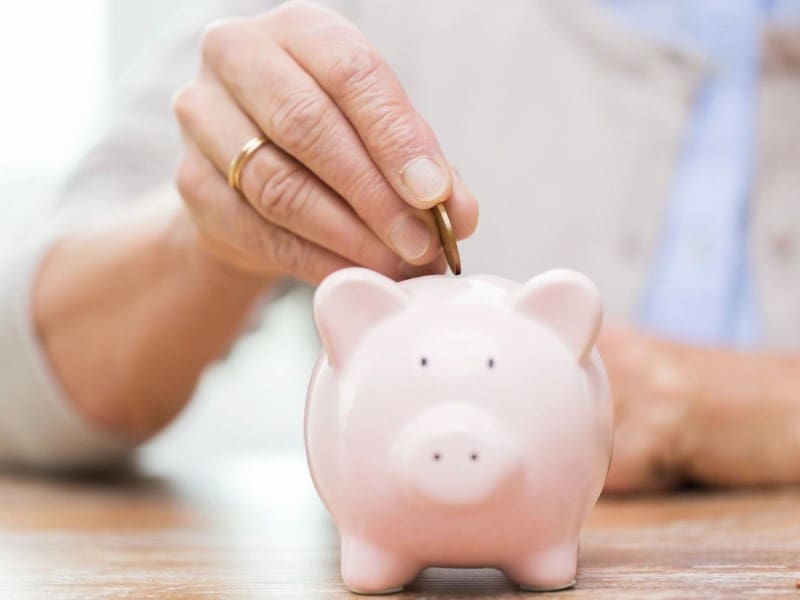A hand is placing a coin into a pink piggy bank on a wooden table.